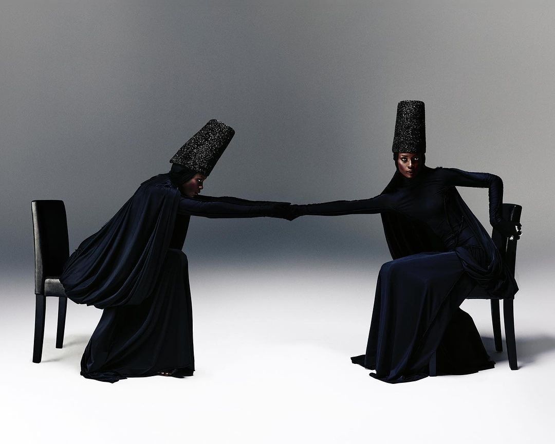 Two figures in black reaching across chairs