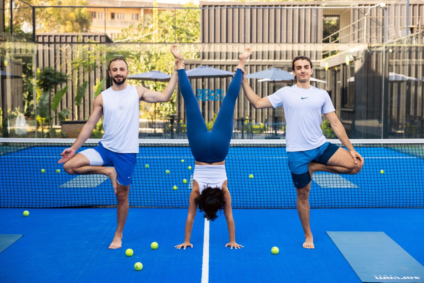 Two teachers in tree pose holding a handstand between them on the padel court